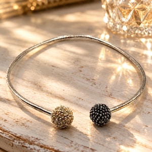 May include: A silver-toned open cuff necklace featuring a textured band and two spherical terminals. One terminal is encrusted with clear, light-reflecting crystals, while the other is covered in small, dark, textured beads for contrast.