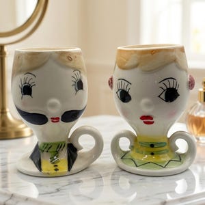 May include: Two ceramic egg cups shaped like human heads. The left cup features a figure with a black mustache, yellow vest, and black jacket. The right cup features a figure with blonde hair, red lips, and a yellow and green patterned top.