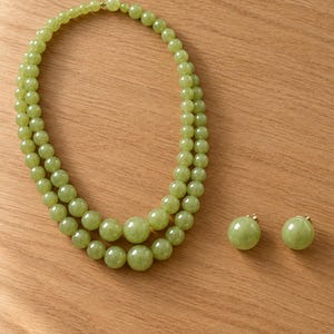 May include: A double-strand necklace featuring smooth, round, light green beads that graduate in size toward the center, paired with a matching set of two round, light green bead stud earrings with gold-toned metal backings.