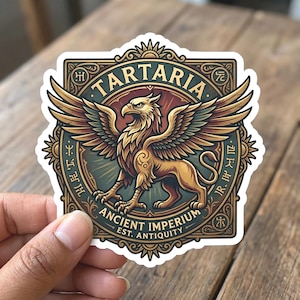 May include: A sticker featuring a golden griffin with outstretched wings, set against a circular background with the text "TARTARIA" and "ANCIENT IMPERIUM EST. ANTIQUITY". The design incorporates ornate borders and sunburst details, with a vintage aesthetic.