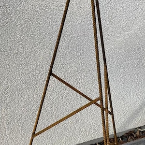May include: A tall, triangular, metal garden trellis with a decorative finial. The trellis is made of brown metal rods and is approximately 150 cm tall. It stands on a concrete surface.