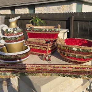 Pode incluir: Uma coleção de itens decorativos, incluindo um jarro e uma tigela dourados, caixas de veludo vermelho e uma cesta redonda. Os itens são adornados com intrincados trabalhos de contas, lantejoulas e franjas em tons de vermelho, dourado e verde.