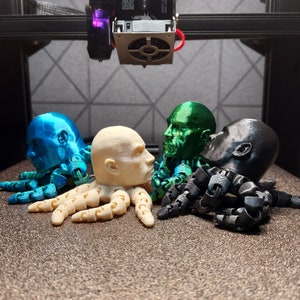 May include: Four 3D printed figurines of human heads, each with a different colour and design. The figurines are arranged in a row, with the first two figurines being blue and green, and the last two figurines being white and black.