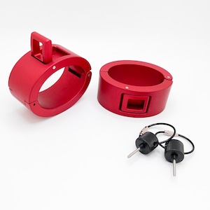 May include: Two red metal cuffs with a locking mechanism and a handle. The cuffs are open, revealing the interior. Two black keys with a silver pin are next to the cuffs. The items are on a white background.