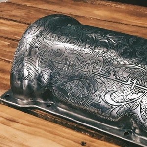 以下が含まれることがあります： 表面に複雑な花柄がエッチングされた銀色のエンジンバルブカバー。カバーの上部に小さな丸い開口部があります。