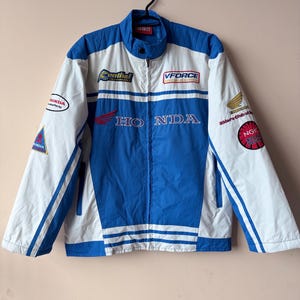 Könnte beinhalten: Eine blaue und weiße Rennjacke mit dem Wort "HONDA" in großen roten Buchstaben auf der Vorderseite. Die Jacke hat mehrere Sponsorenlogos, darunter "VFORCE", "Central" und "NGK SPARK PLUGS".