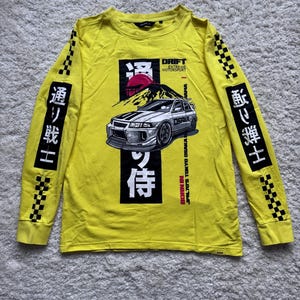 Op de afbeelding: Geel shirt met lange mouwen met een afbeelding van een auto, een berg en Japanse tekst. Het shirt heeft de tekst "DRIFT MOTORSPORT" en "BORN IN JPN.70'S TOKYO OSAKA". Geruite patronen staan op de mouwen.