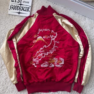 May include: Red satin bomber jacket with gold sleeves and a map of Japan embroidered on the back. The jacket features the word "Japan" in script and various city names. The sleeves are a light gold colour.