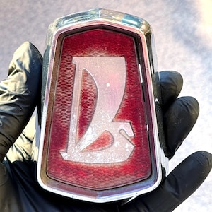 Puede incluir: Emblema de coche vintage con marco cromado y fondo rojo. El emblema presenta un diseño estilizado en plata de la letra "L". El emblema es rectangular con la parte superior curvada y está sujeto por una mano enguantada.