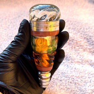 May include: A decorative bottle stopper with a clear, textured top. The stopper features a silver band, a brown band, and a section with a yellow rose design. The bottom is a silver base.