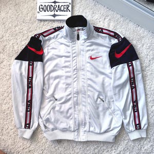 May include: White track jacket with black and red accents. Features a red Nike swoosh logo on the chest and "USA" printed on the sleeves. The jacket has a full zipper and two front pockets. The Etsy store name "GOODRACER" is visible in the background.