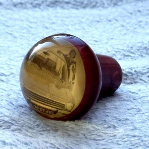 Op de afbeelding: Een paar vintage-stijl, rode en gouden glazen cabochons. Eén cabochon heeft een sepia-getinte afbeelding van een persoon naast een auto, met de nummerplaat "06-DT-69".