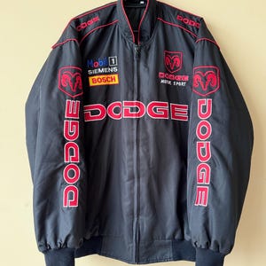 Puede incluir: Chaqueta negra con logotipos bordados en rojo y blanco. Los logotipos incluyen "Dodge", "Mobil 1", "Siemens", "Bosch" y "Dodge Motor Sport".