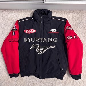 Op de afbeelding: Zwarte en rode Mustang jas met het Mustang logo en de tekst "40th Anniversary". Voorzien van het Ford logo en "5.0" op de mouwen. De jas heeft een bomberstijl met een knoopsluiting.