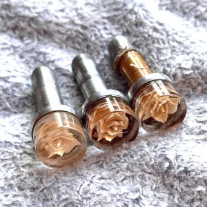 Op de afbeelding: Drie decoratieve flessenstoppers met een heldere harsbasis, elk met een delicate, crèmekleurige roos. De stoppers hebben zilverkleurige metalen accenten en een cilindrische vorm, voor wijnflessen.