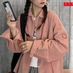 Puede incluir: Camisa de pana rosa claro con botones, bolsillo en el pecho y mangas largas. La camisa tiene una pequeña carita sonriente bordada en la manga izquierda. Parte de una colección de ropa casual.