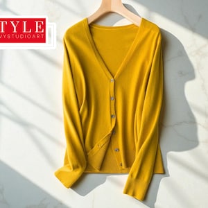 May include: Mustard yellow cardigan sweater with a V-neck and button closure. The long-sleeved sweater is hanging on a wooden hanger. The text "STYLE SAVVYSTUDIOART" is in a red and white rectangle in the upper left corner.
