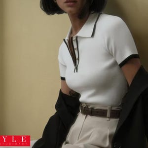 May include: A white polo shirt with black trim and a black jacket. The shirt has a ribbed texture and a collar. The outfit includes beige trousers and a brown belt. The image has the text "STYLE SAVVYSTUDIOART" in the bottom left corner.