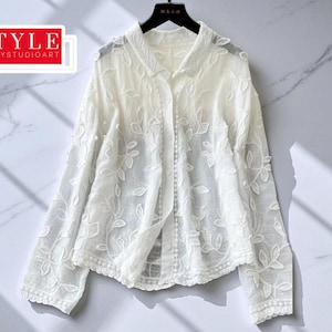 May include: A white, embroidered blouse with a floral design. The long-sleeved top has a button-down front and a collar. The sheer fabric is visible. The blouse hangs on a black hanger against a marble backdrop. The text "STYLE SAVVYSTUDIOART" is in a red box.