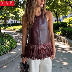 Puede incluir: Un top de cuero burdeos sin mangas con dobladillo de flecos. El top se combina con pantalones blancos. La imagen tiene el texto "STYLE SAVVYSTUDIOART". El fondo es un parque con árboles y vegetación.
