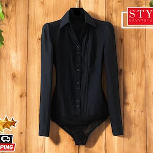 Puede incluir: Una camisa negra de manga larga con botones en la parte delantera y un detalle plisado en la parte delantera. La camisa tiene un cuerpo ajustado y un dobladillo ligeramente acampanado.