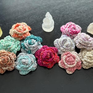 May include: A selection of handmade crochet rose flowers in various colours, including orange, teal, pink, and beige. The roses are arranged with crystals and a small, clear sculpture on a dark surface.