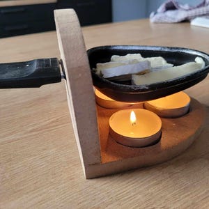 Könnte beinhalten: Ein Käsefondue-Set aus Holz mit einem schwarzen Topf aus Gusseisen und drei brennenden Teelichtern. Der Topf ist mit Käse und Brot gefüllt.