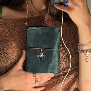 May include: A dark teal leather journal with a smooth, matte finish and a vertical elastic band closure. A silver-toned starburst charm with a small central blue stone is attached to the elastic band, resting against the front cover of the journal.