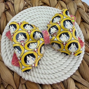 May include: A hair bow featuring a repeating pattern of the anime character Monkey D. Luffy wearing a yellow straw hat. The bow has a central band and side accents made of pink glitter material, with the main sections in a smooth white finish.