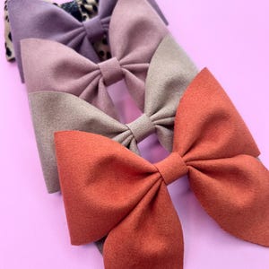May include: Three fabric hair bows in different colors and patterns. The top bow is a leopard print with a purple bow underneath. The middle bow is a light brown with a pink bow underneath. The bottom bow is a solid orange.