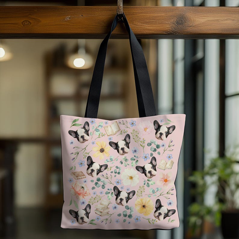 May include: A pink tote bag with a floral pattern featuring a repeated image of a French bulldog. The bag has black straps and a white background with a variety of flowers, leaves, and books.