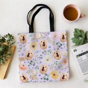 May include: A lavender tote bag with a black handle features a repeating pattern of golden retriever dog faces, flowers, and greenery.