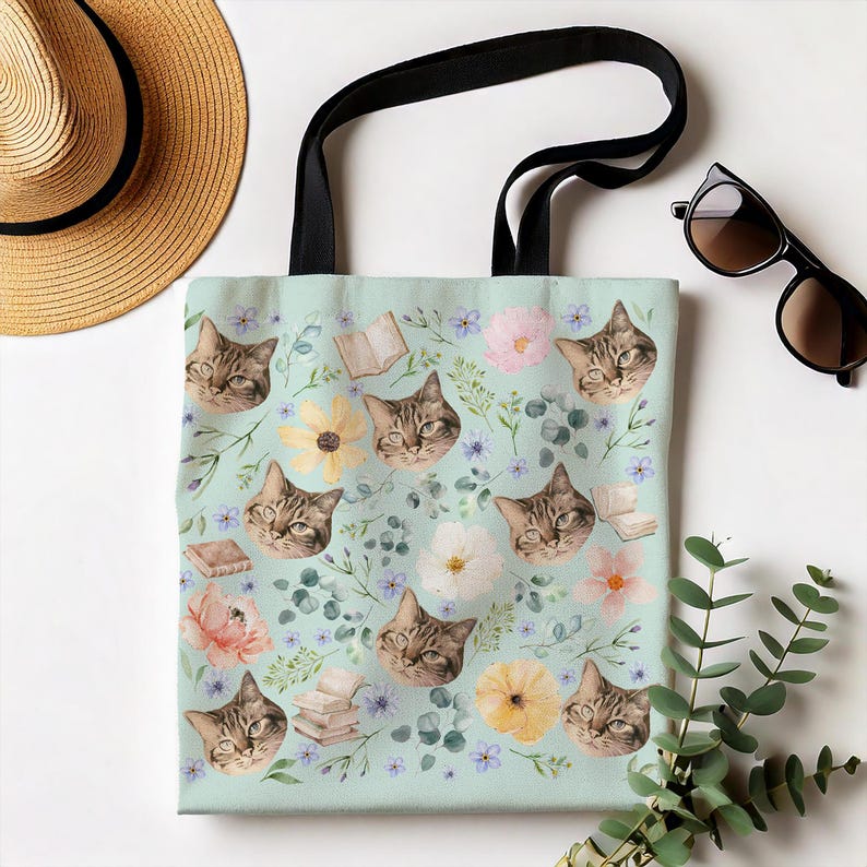 May include: A light blue tote bag with a repeating pattern of brown tabby cats, flowers, and books. The bag has black straps.