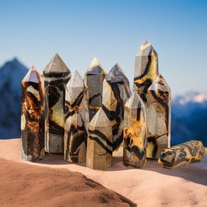 Puede incluir: Un conjunto de cristales pulidos en forma de torre, con una mezcla de colores marrón, negro y crema. Los cristales tienen facetas geométricas y patrones variados. El fondo muestra un cielo azul y una cordillera.