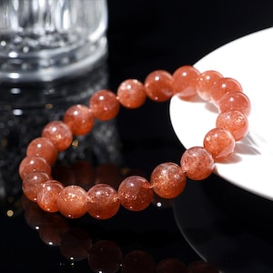 Op de afbeelding: Een armband met ronde, rood-oranje zonnesteen kralen. De kralen hebben een gespikkelde uitstraling. De armband wordt gepresenteerd op een wit oppervlak tegen een donkere achtergrond. Elke kraal is ongeveer 1 cm in diameter.