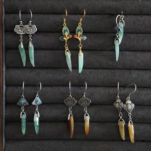 Op de afbeelding: Een verzameling handgemaakte oorbellen, tentoongesteld op een donkergrijze ondergrond. De oorbellen hebben verschillende ontwerpen, waaronder zilveren filigraan, turkooizen accenten en jade- of amberkleurige hangers. Sommige hebben goudkleurige accenten.