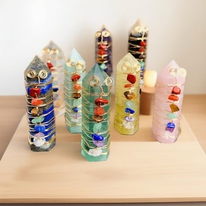 May include: Seven crystal wands with gold wire wrapped around them, each with different colored gemstones. The wands are arranged on a wooden surface.