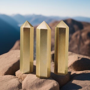 May include: Three gold-colored, hexagonal-shaped, crystal-like objects stand upright on a rocky surface. The objects are arranged in a row, with the middle object slightly taller than the others.