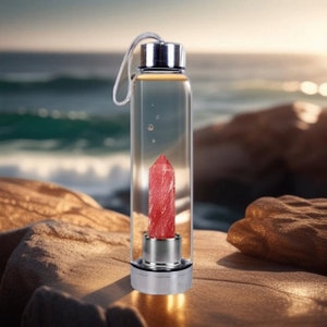 May include: A clear glass water bottle with a stainless steel lid and a red crystal inside. The bottle is sitting on a sandy beach.