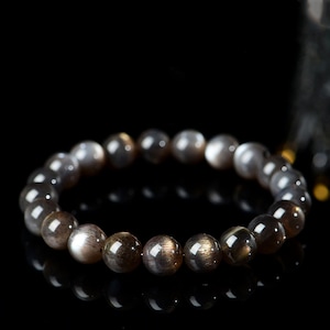 Puede incluir: Un brazalete de cuentas de piedra de sol negra con un brillo plateado. Las cuentas son redondas y lisas, y el brazalete está hecho de piedra natural.
