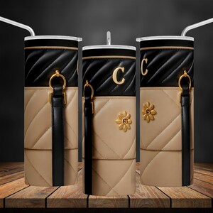 Puede incluir: Tres vasos de acero inoxidable con diseños acolchados en negro y beige. Los vasos tienen una correa negra con un anillo dorado y un acento de flor dorada. Los vasos están personalizados con la letra "C".