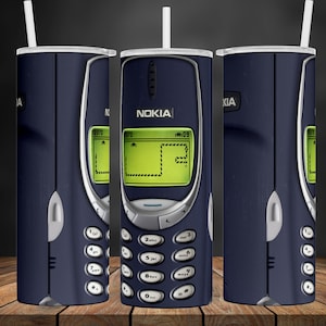 May include: A navy blue tumbler featuring a retro Nokia phone design. The design includes the Nokia logo, a green screen displaying a game, and a keypad. The tumbler has a white straw and a silver rim.