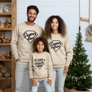 以下が含まれることがあります： 3人家族が、黒い文字が入ったベージュのトレーナーを着ています。大人のトレーナーには、ダイヤモンド型の「スーパーパパ」と「スーパーママ」の文字が書かれています。子供のトレーナーには、ダイヤモンド型の「ママとパパが大好き」の文字が書かれています。