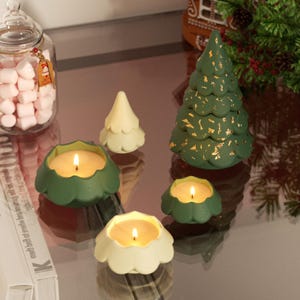 May include: A selection of festive candles and decorations. The image shows green and cream-coloured tree-shaped candles, alongside candles in green and cream-coloured floral-shaped holders. A large green tree candle with gold accents is also visible.