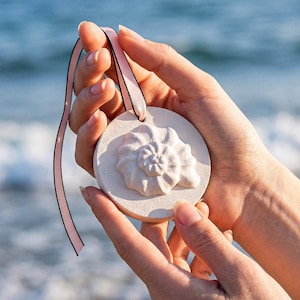 Op de afbeelding: Een ronde, gebroken witte schelpenornament met een gedetailleerd spiraalontwerp. Het ornament wordt vastgehouden door een roze lint en door handen. De achtergrond is een wazig uitzicht op de zee.