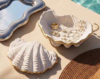Op de afbeelding: Een verzameling decoratieve trays en sieraden. Een zilverkleurige, golvende spiegeltray staat naast een witte, schelpvormige schaal met gouden rand, met ringen en kettingen. Een tweede schelpvormige tray is ook aanwezig. Een strohoed staat in de rechter benedenhoek.