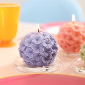 May include: A lavender hydrangea-shaped candle with a lit wick sits on a clear glass plate. The candle is surrounded by other spherical candles in shades of pink and green. A yellow glass is in the background.