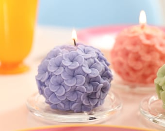 May include: A lavender hydrangea-shaped candle with a lit wick sits on a clear glass plate. The candle is surrounded by other spherical candles in shades of pink and green. A yellow glass is in the background.