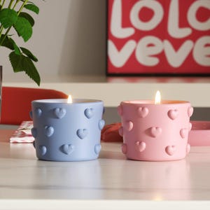 Könnte beinhalten: Zwei herzförmige Kerzen, eine blaue und eine rosa, mit einem erhabenen Herzmuster. Die Kerzen stehen auf einer weißen Oberfläche.
