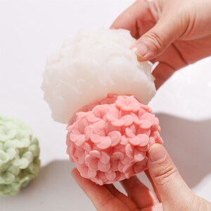 May include: A close-up of a pink floral-shaped object being held, with a white floral-shaped object above it. A green floral-shaped object is in the background. The objects appear to be molds for soap or other crafts.